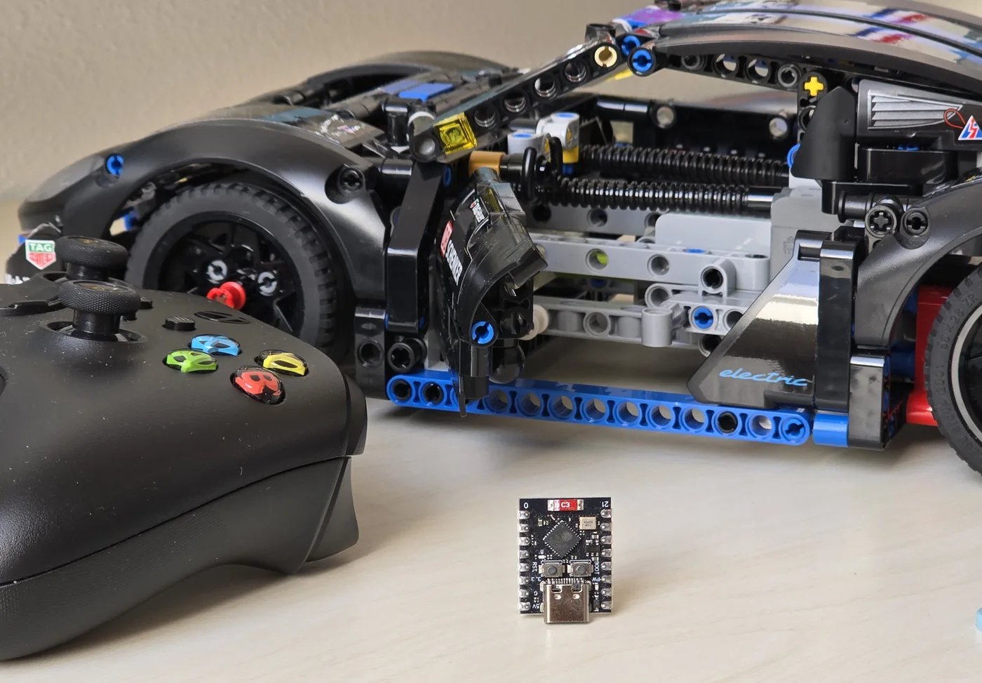 Nahaufnahme der ESP32-C3-Platine mit USB-C-Buchse auf dem Schreibtisch, dahinter unscharf der Xbox-Controller und das Fahrzeug.